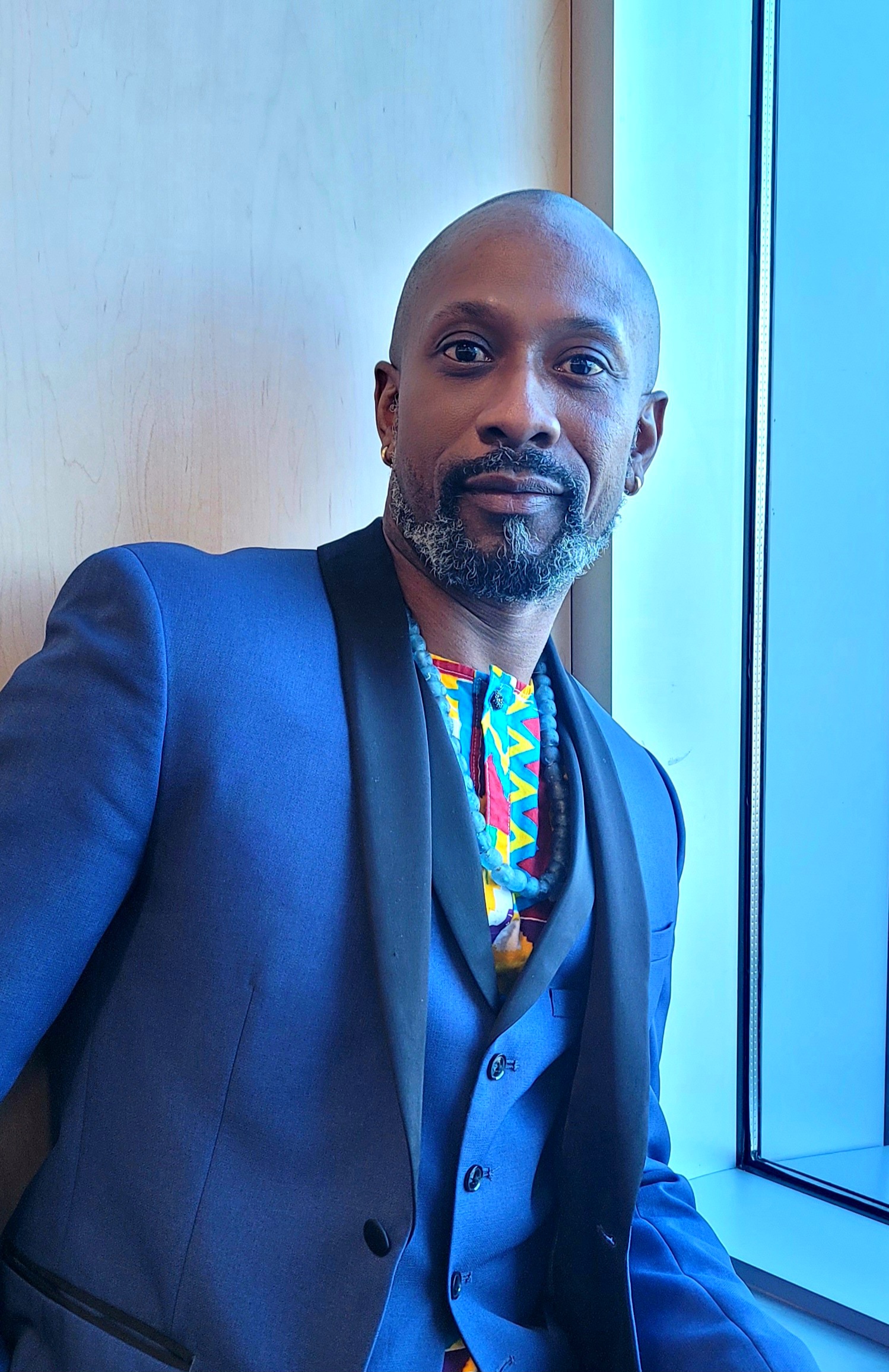 A photo of a man in a blue suit, colourful shirt and light blue beads