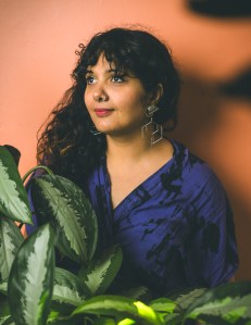 Salima, a brown skinned woman with long curly dark hair stares into the distance. She is surrounded by plants contrasted against a peachy pink wall.